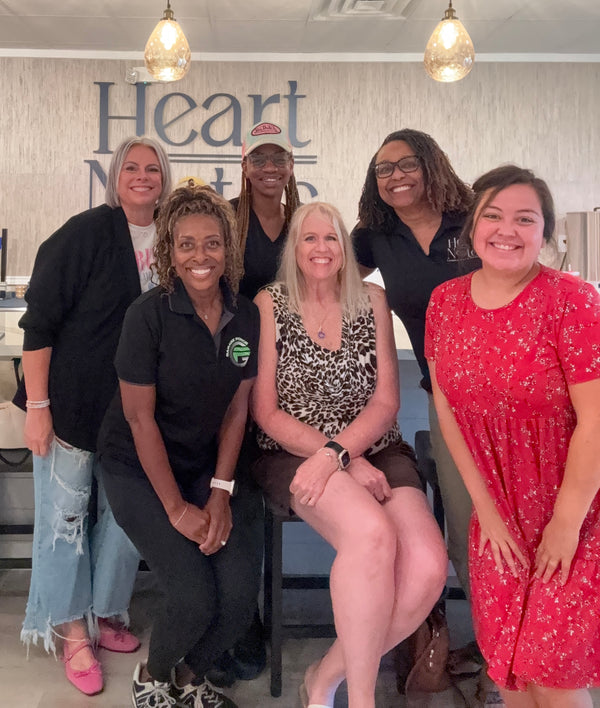 Six women smiling and posing together in a bright indoor space with a wall sign reading 'Heart Notes' behind them.